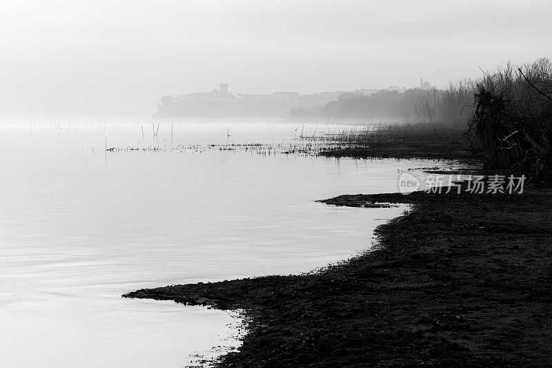 黎明时分，美丽的trasiimeno湖(翁布里亚)岸边，背景是薄雾中的Castiglione del Lago镇